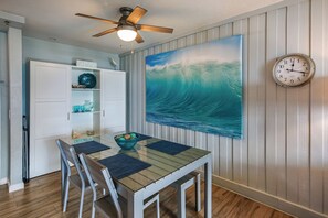A dining area with a glass table, chairs, a ceiling fan, a clock, and a large wave painting on the wall.