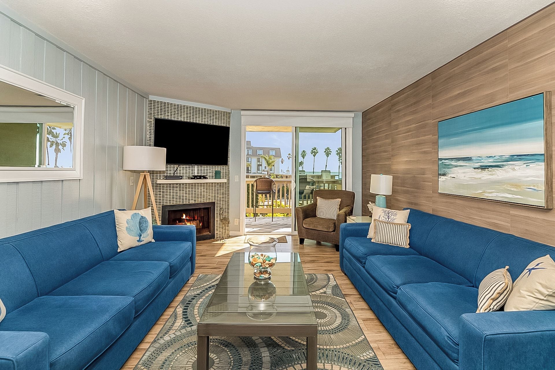 A living room with two blue sofas, a glass coffee table, a fireplace, and a large window with a view of palm trees.