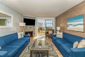 A living room with two blue sofas, a glass coffee table, a fireplace, and a large window with a view of palm trees.