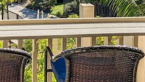 A balcony with wicker chairs, a view of the ocean, palm trees, and a building with a pool.
