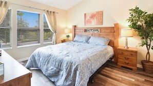 A bedroom with a large bed, a nightstand, a lamp, a potted plant, and a window with curtains.