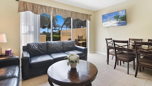 A living room with a leather sofa, a dining table with chairs, a TV on the wall, and a view of trees and sky through the window.