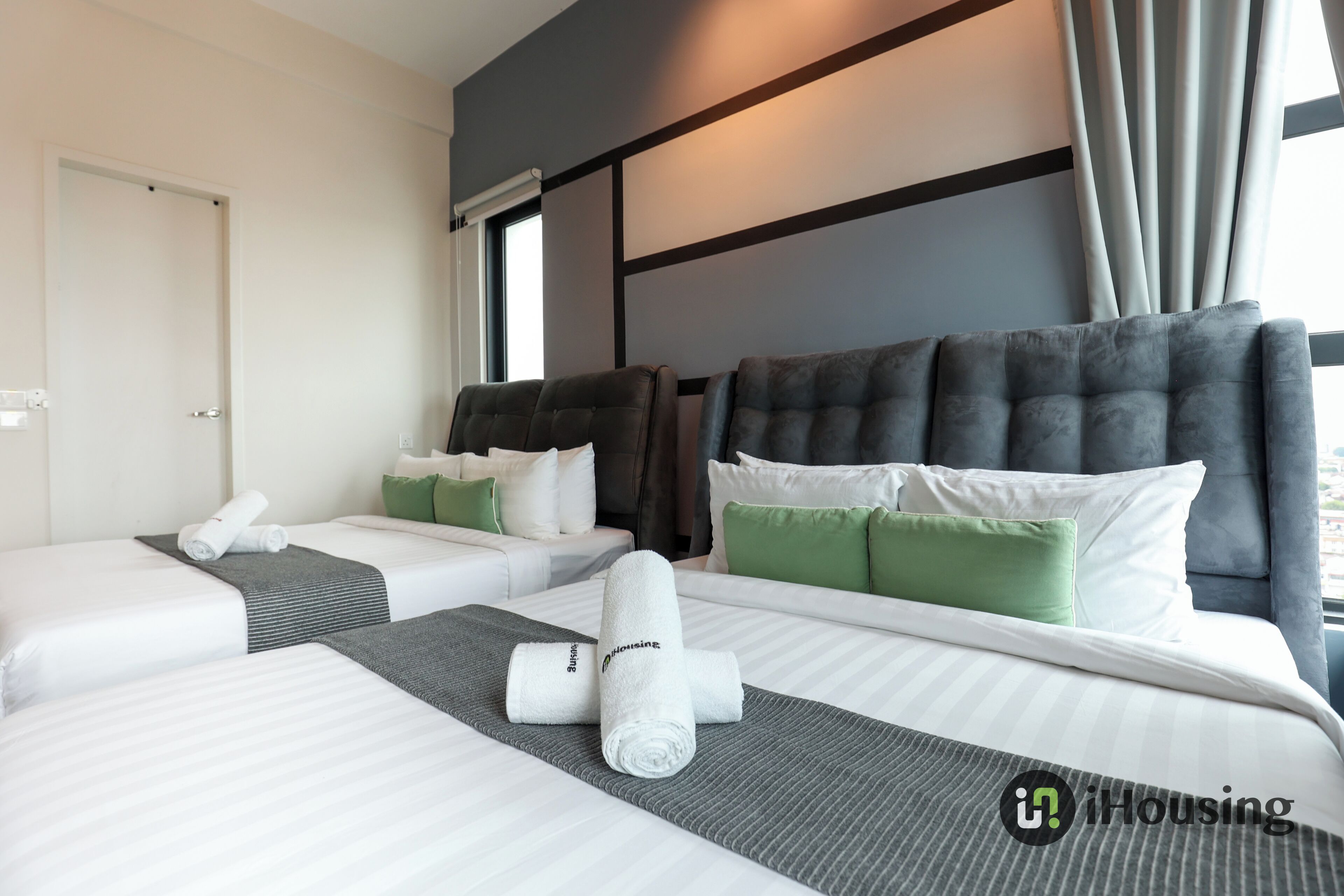 A hotel room with two beds, grey and white bedding, green and white pillows, and a window with curtains.