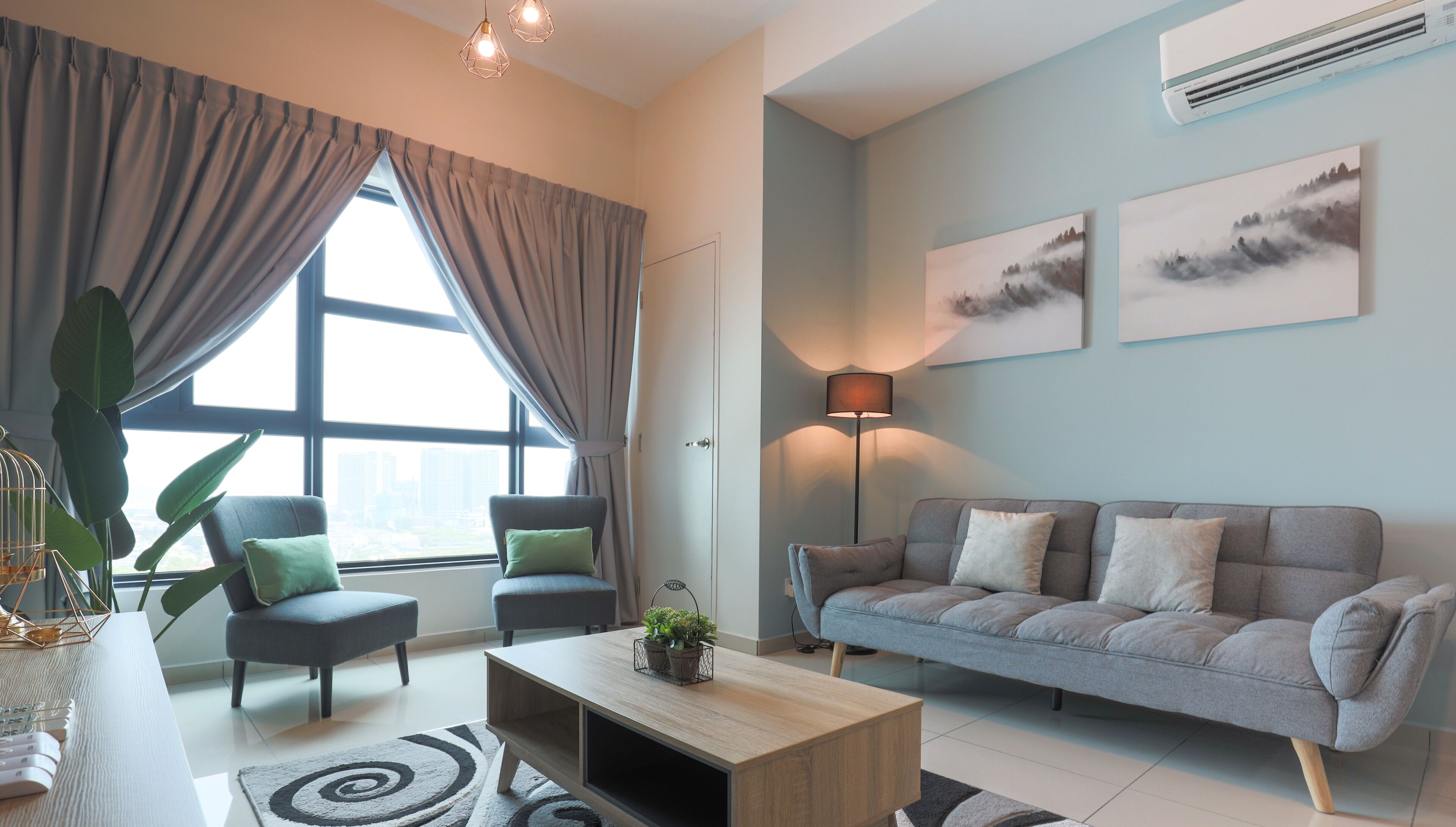 A modern living room with a grey sofa, two armchairs, a coffee table, and a large window with curtains. There are two wall-mounted air conditioning units and two framed pictures on the wall.
