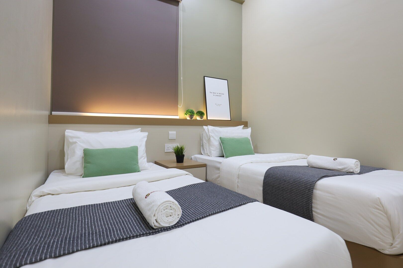 A hotel room with two beds, each with white linens and green pillows, a small bedside table with a plant, and a framed inspirational quote on the wall.