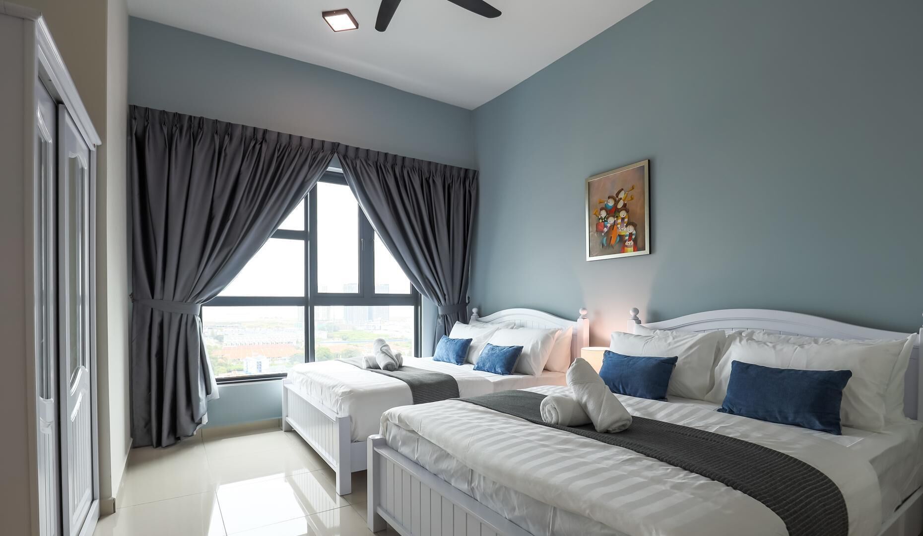 Una camera d'albergo con due letti, una ventola a soffitto, una finestra con tende e un quadro appeso al muro.