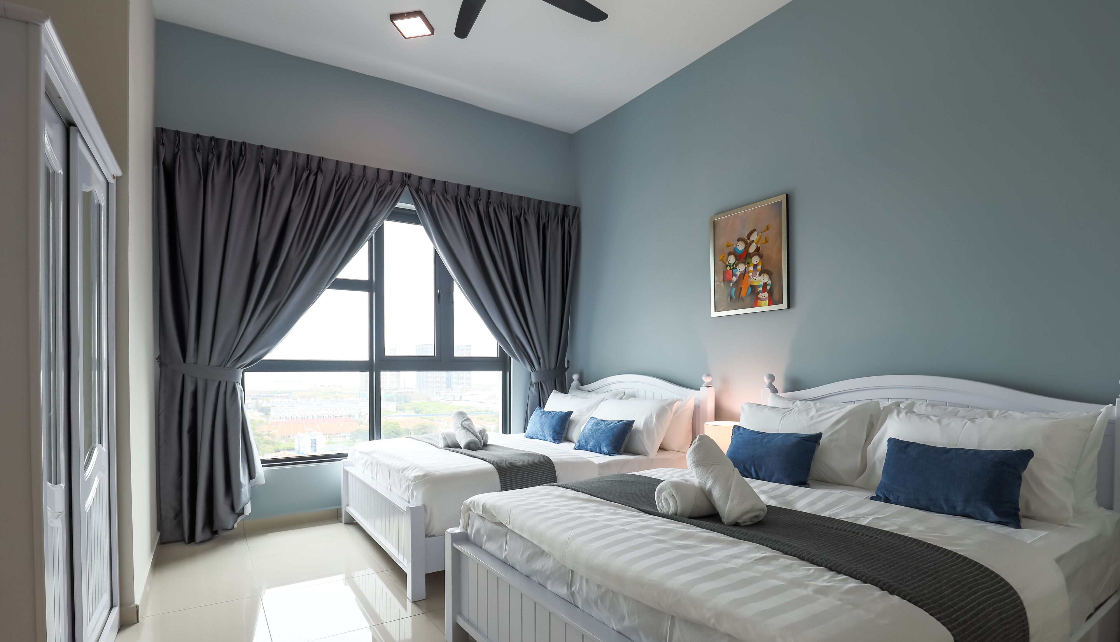 Une chambre moderne avec deux lits, une grande fenêtre avec des rideaux, un ventilateur de plafond et un tableau au mur.