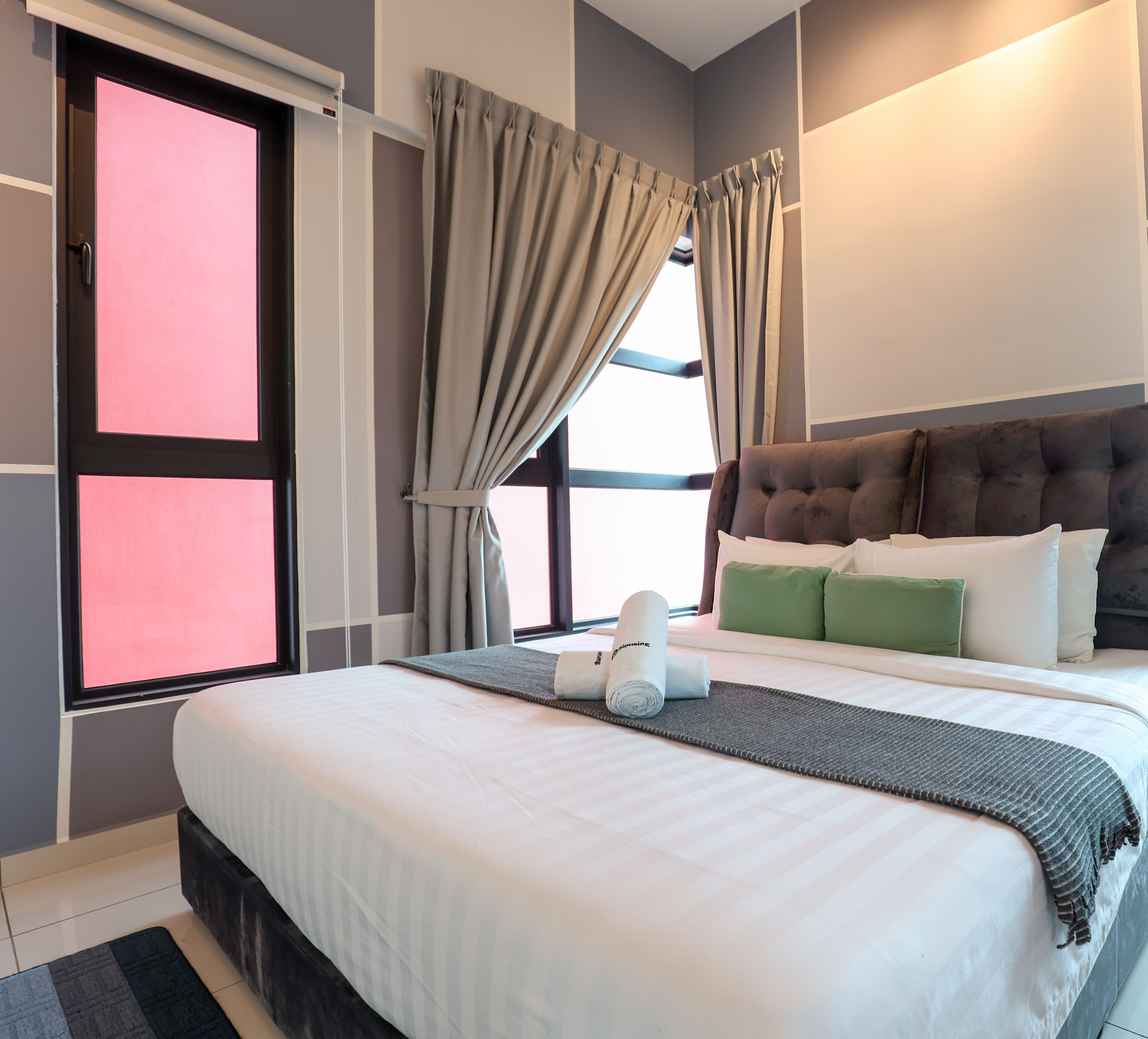 A modern hotel room with a large bed, grey curtains, a tufted headboard, and a view of the outdoors.