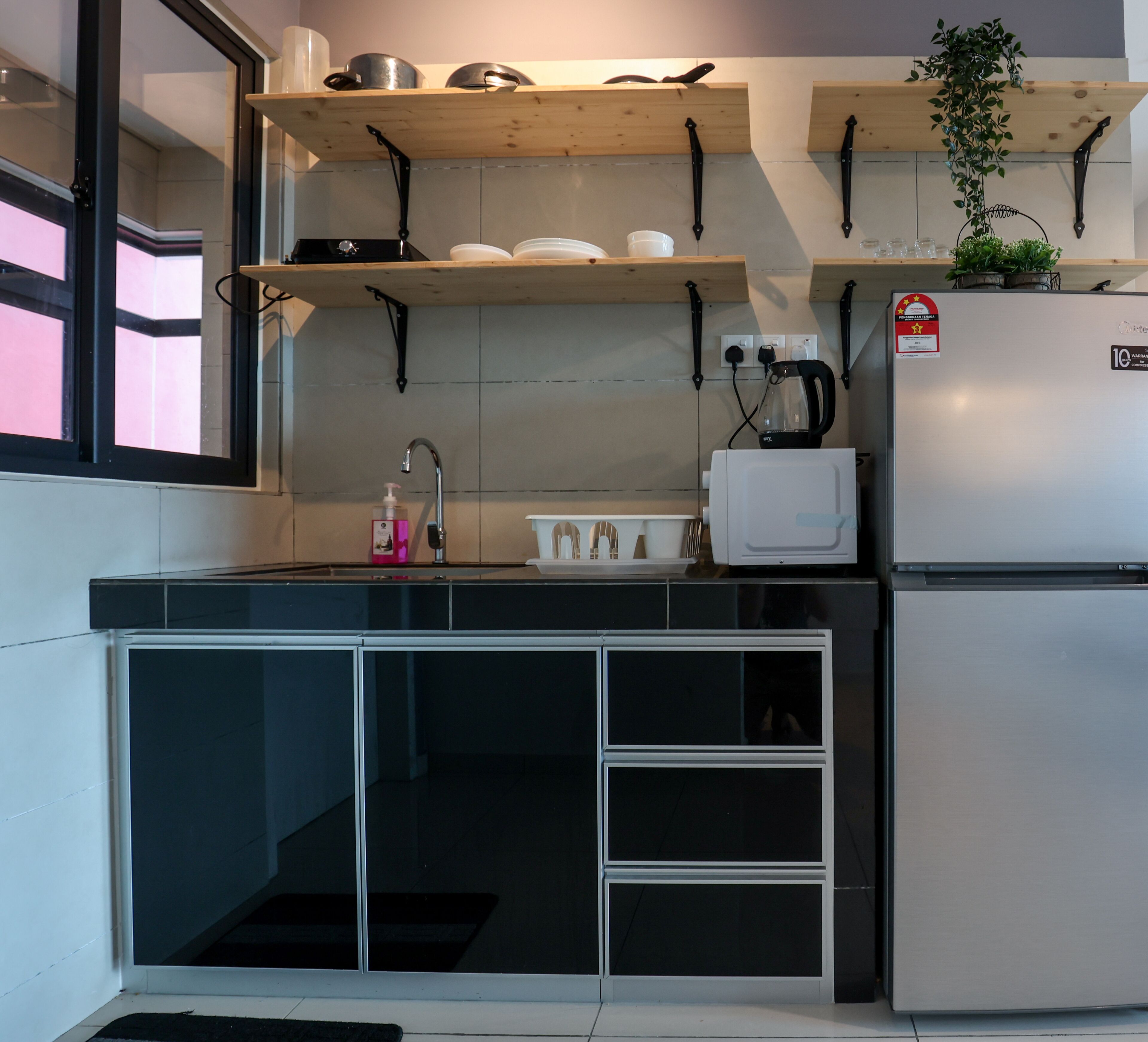 Een compacte keuken met een ingebouwde kookplaat, een spoelbak en een koelkast. Boven de kookplaat bevinden zich planken met pannen en een potplant.