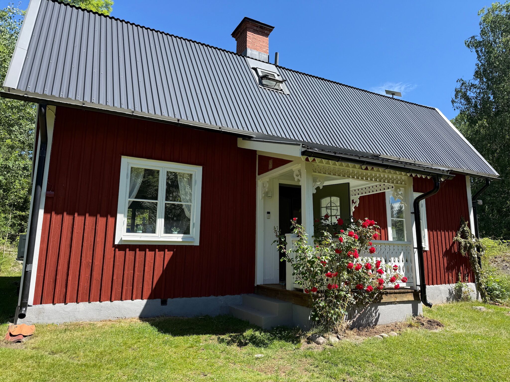 Ett rött trähus med ett grått tak, en vit veranda och en liten trädgård med röda blommor.