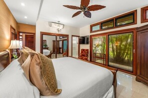 Une chambre spacieuse avec un grand lit, un ventilateur de plafond et une vue sur la verdure par les fenêtres.