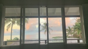 Una habitación con un amplio ventanal con vista a una playa, palmeras y un atardecer.