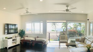 Un salón amplio con televisor grande, asientos cómodos, ventilador de techo y un balcón con vista a palmeras.