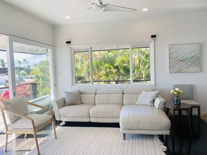 A modern living room with a beige sectional sofa, a round wooden side table with a vase of flowers, and a large window offering a view of lush greenery.