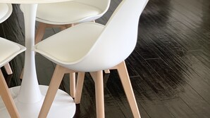 Un comedor moderno con una mesa redonda blanca, sillas blancas con patas de madera y un jarrón azul con flores blancas.