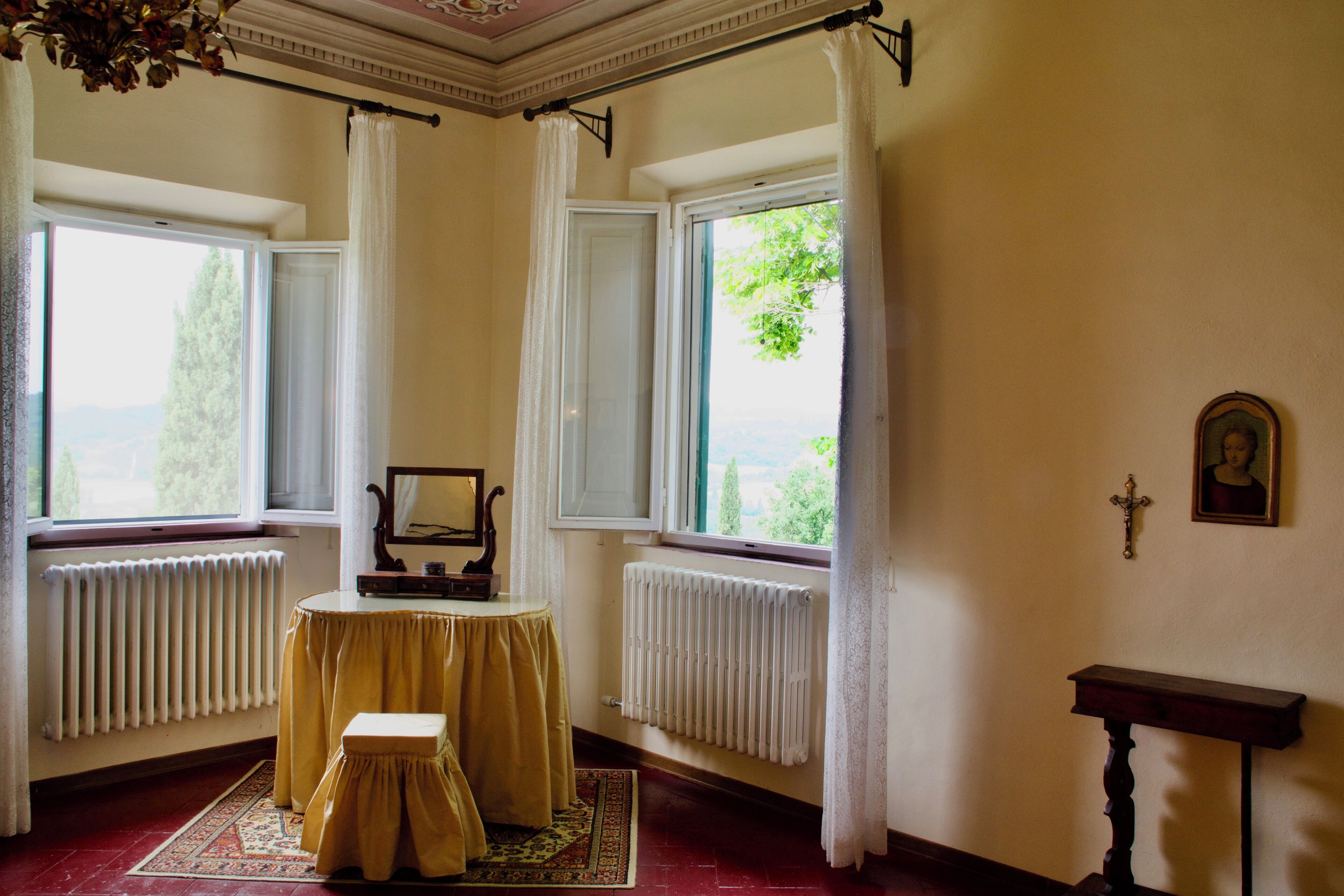 A room with two windows, a table with a chair, a radiator, and a small wooden stand with a cross.