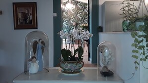 Huoneessa on valkoinen pöytä, jonka päällä on maljakko valkoisia orkideoita, koristeellinen kulho ja lasiveistos. Seinällä on kehystetty kuva ja peili, joka heijastaa oviaukkoa.