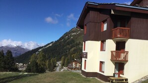 Ein zweigeschossiges Gebäude mit Balkon, umgeben von einer Rasenfläche mit Schneeflecken, vor einer Bergkulisse.