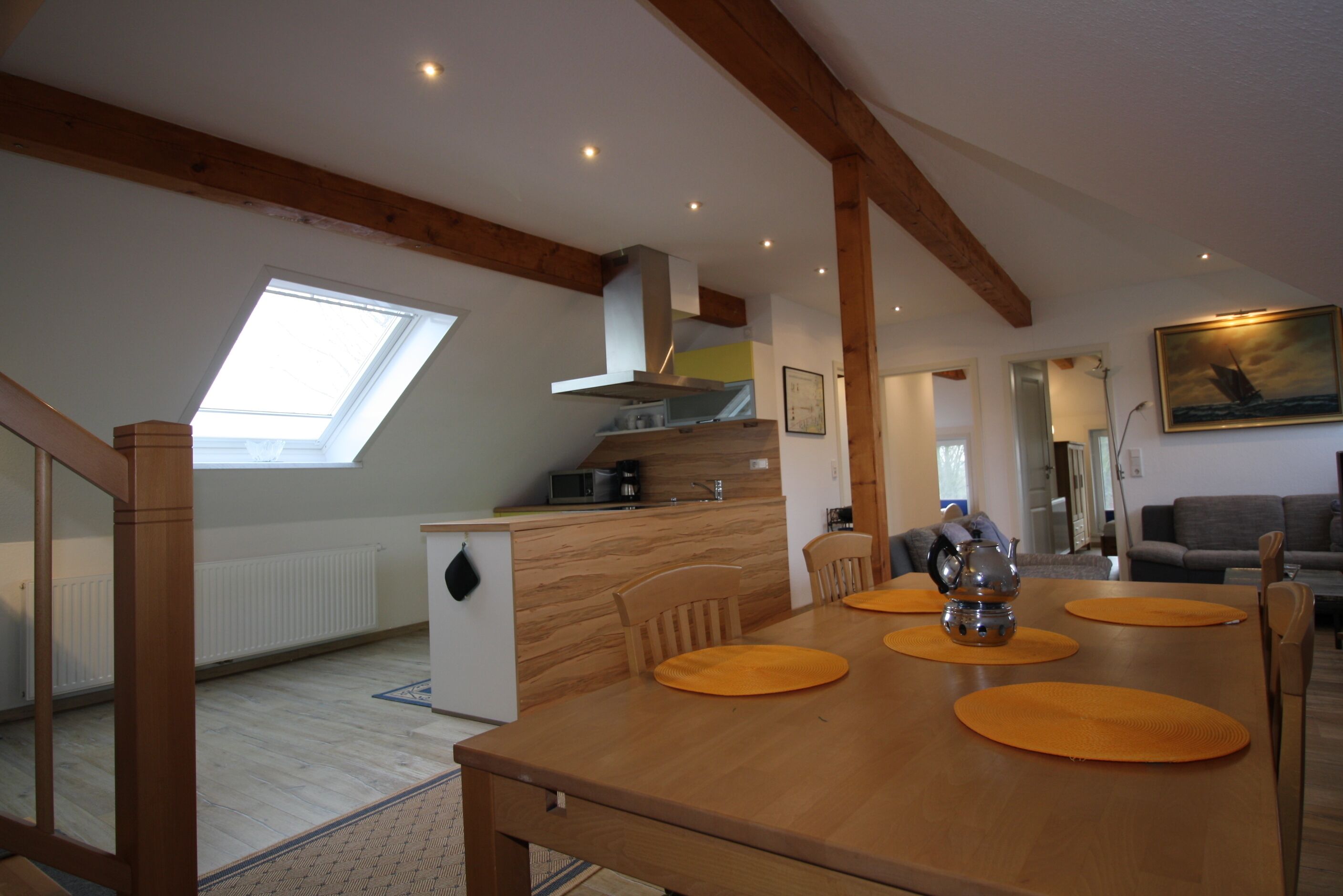 A dining area with a wooden table, orange placemats, and a teapot. There is a kitchen in the background with a microwave and a coffee maker.