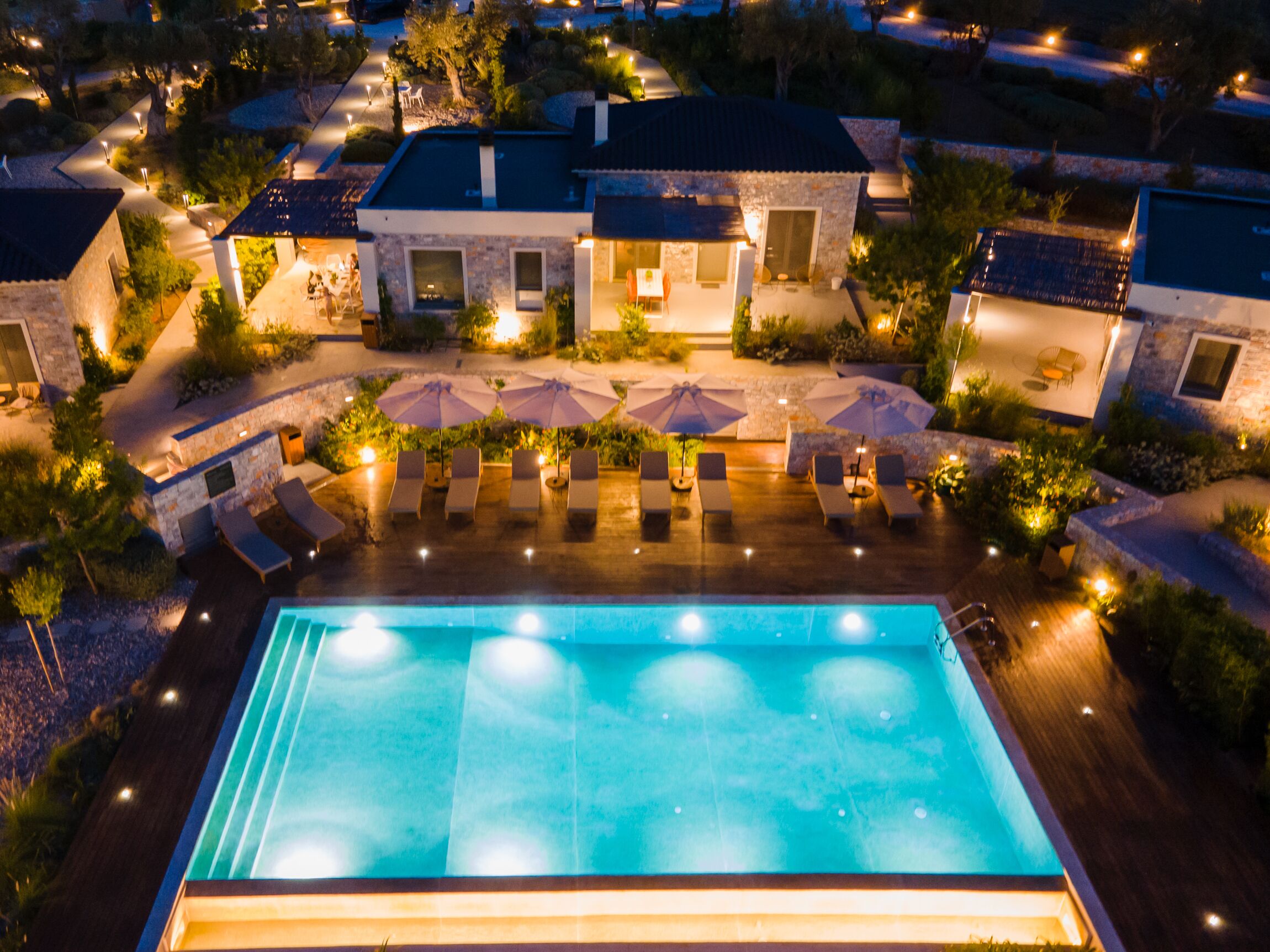 A swimming pool with lounge chairs and umbrellas, surrounded by stone buildings and well-lit pathways.