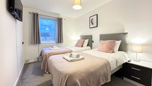 A hotel room with two beds, a TV mounted on the wall, a window with curtains, a bedside lamp, and a framed picture on the wall.