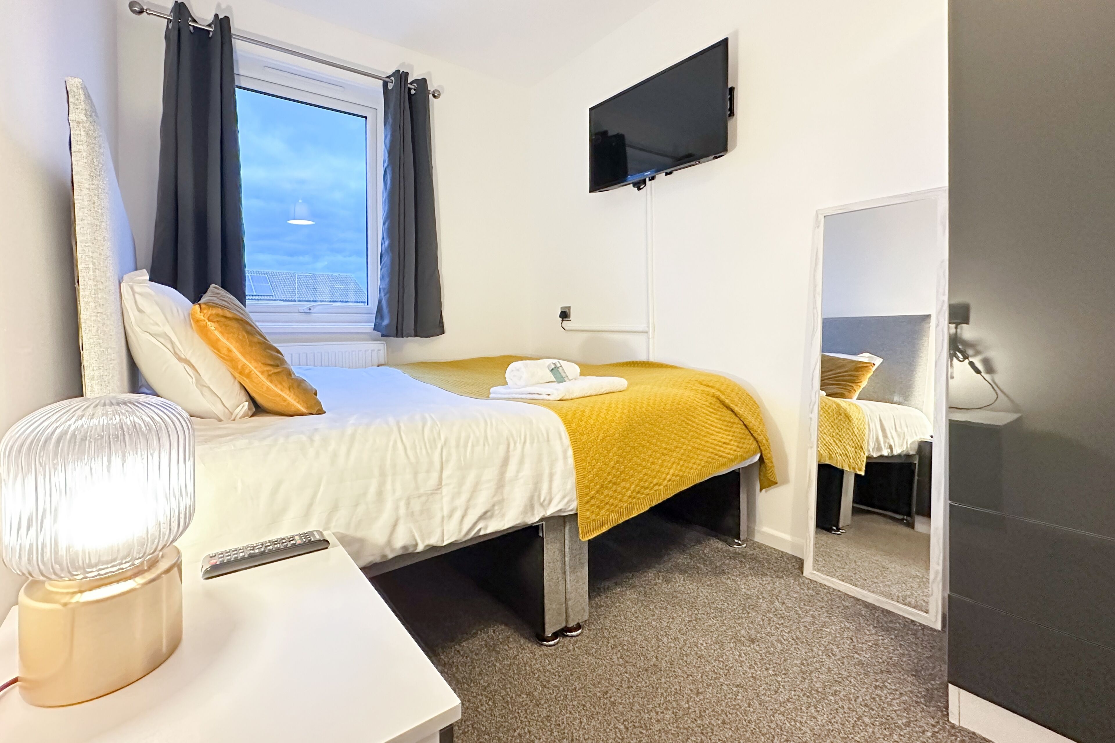 A hotel room with a bed, a TV, a bedside table with a lamp, and a window with curtains.