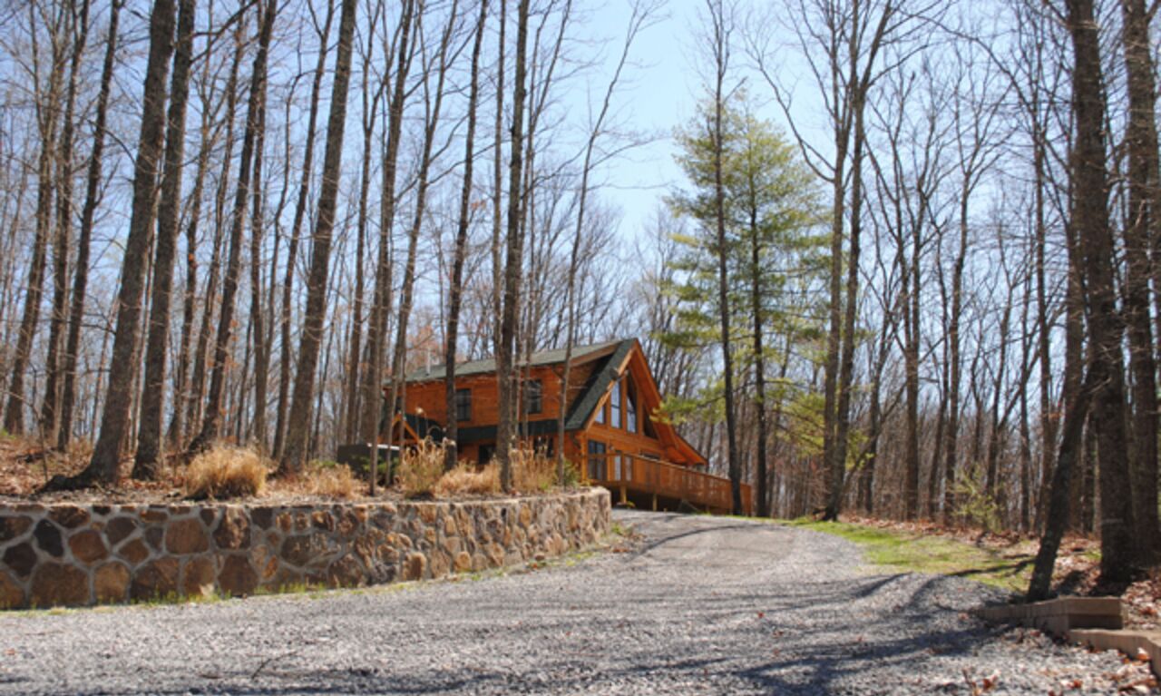 Une cabane avec un mur de soutènement en pierre et une allée de gravier menant à celle-ci, le tout entouré de grands arbres.