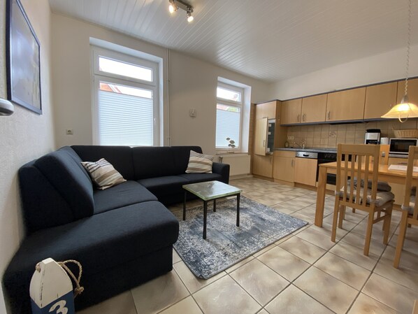 A modern living room with a dark sofa, a small coffee table, and a dining area with wooden chairs and a table. - "Kleine Möwe" Vacation Apartment with a Large Garden in the Heart of Pilsum (Krummhörn)