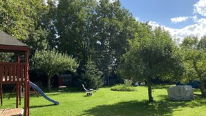 Un patio trasero con césped, una estructura de juego de madera, un tobogán y una zona de estar cubierta.