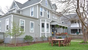 A two-story house with a front porch, a dining area with a table and chairs, and a small garden area.