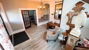 A room with a wooden floor, a beige armchair, a wooden side table, a mirror with a decorative frame, and a chandelier.