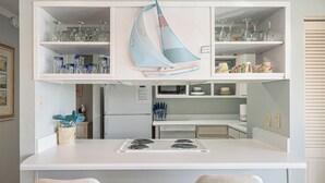 A kitchen with a white countertop, two burners, and a microwave. There are shelves above the counter holding glassware and jars. - Relax near Destin Beach. Destin’s best resort style condo! (Destin)