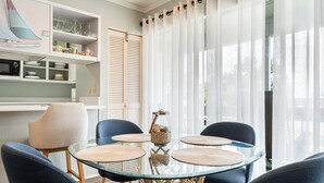 A dining area with a glass table surrounded by chairs, a wicker chair, and a kitchen area with shelves and a microwave. - Relax near Destin Beach. Destin’s best resort style condo! (Destin)