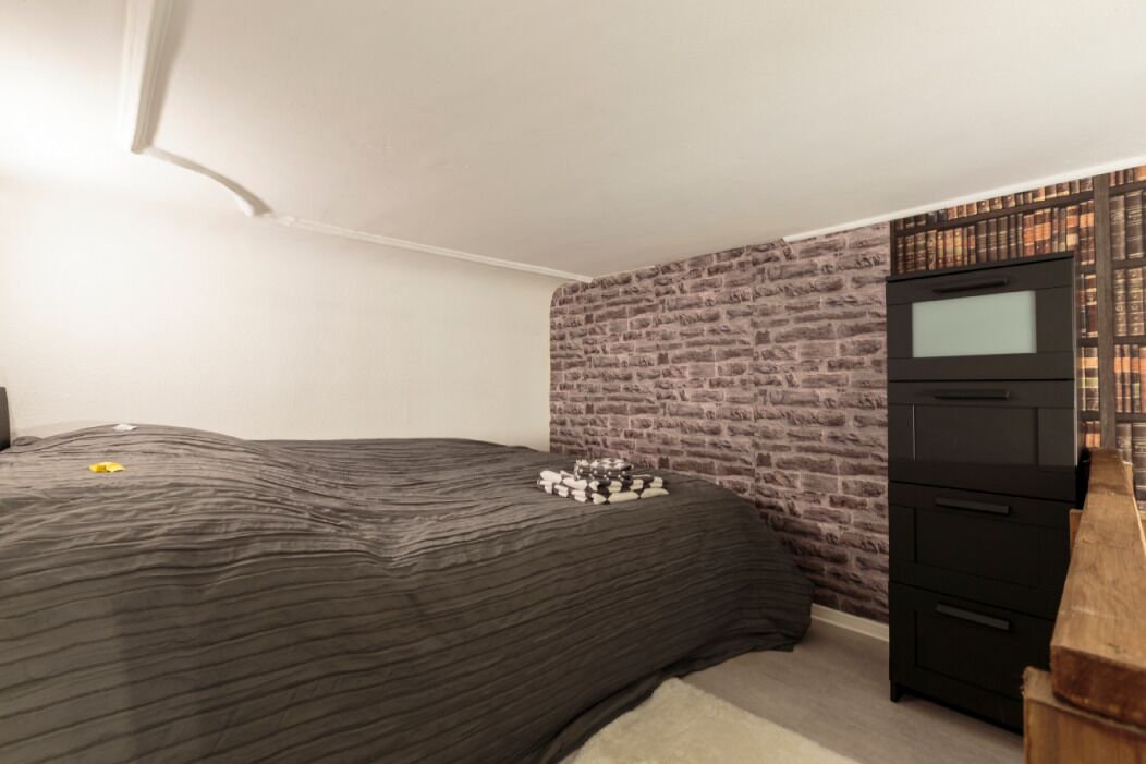 A bedroom with a brick-patterned wall, a bed with a dark striped bedspread, and a black dresser with drawers.