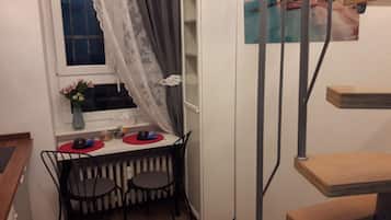 A small dining area with a table and chairs, a radiator, a window with curtains, and a painting on the wall.