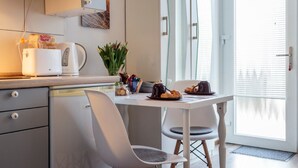 Eine moderne Küche mit weißer Arbeitsplatte, einer Kaffeemaschine, einem Toaster und einer Pflanze. Es gibt einen Esstisch mit Stühlen und ein Fenster mit Jalousien.