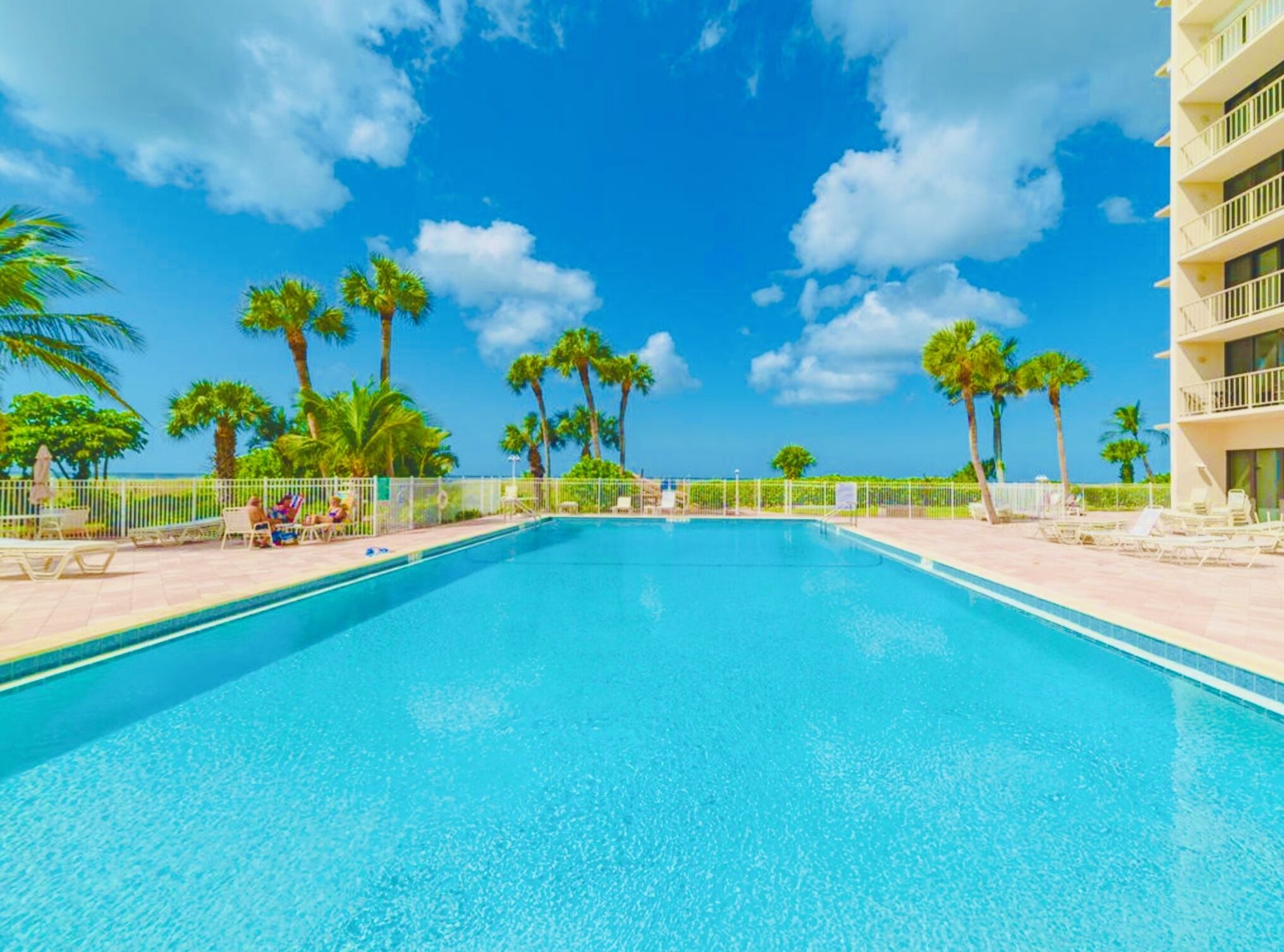 A clear blue pool with lounge chairs and palm trees, surrounded by a fence and a building with balconies.