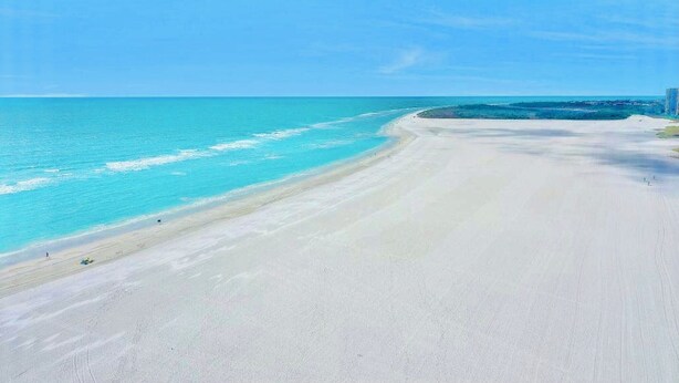 Ein breiter, sandiger Strand mit klarem, türkisblauem Wasser und einem Gebäude in der Ferne rechts.