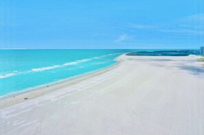 Uma praia extensa e arenosa, com águas turquesa cristalinas e um edifício ao longe, à direita.
