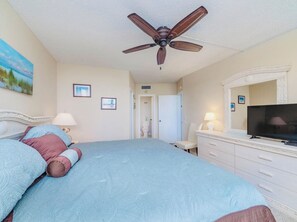 Un dormitorio con cama, ventilador de techo, televisión y puerta de baño al fondo.