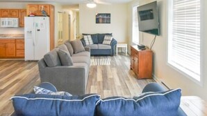 Un salon avec un canapé gris et des coussins bleus, une cuisine avec des meubles en bois et un téléviseur fixé au mur.
