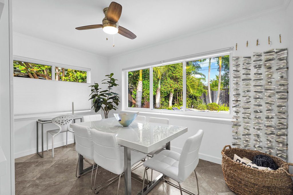 Un espace repas moderne avec une table et des chaises blanches, un ventilateur de plafond, de grandes fenêtres donnant sur un paysage verdoyant et un élément décoratif mural.