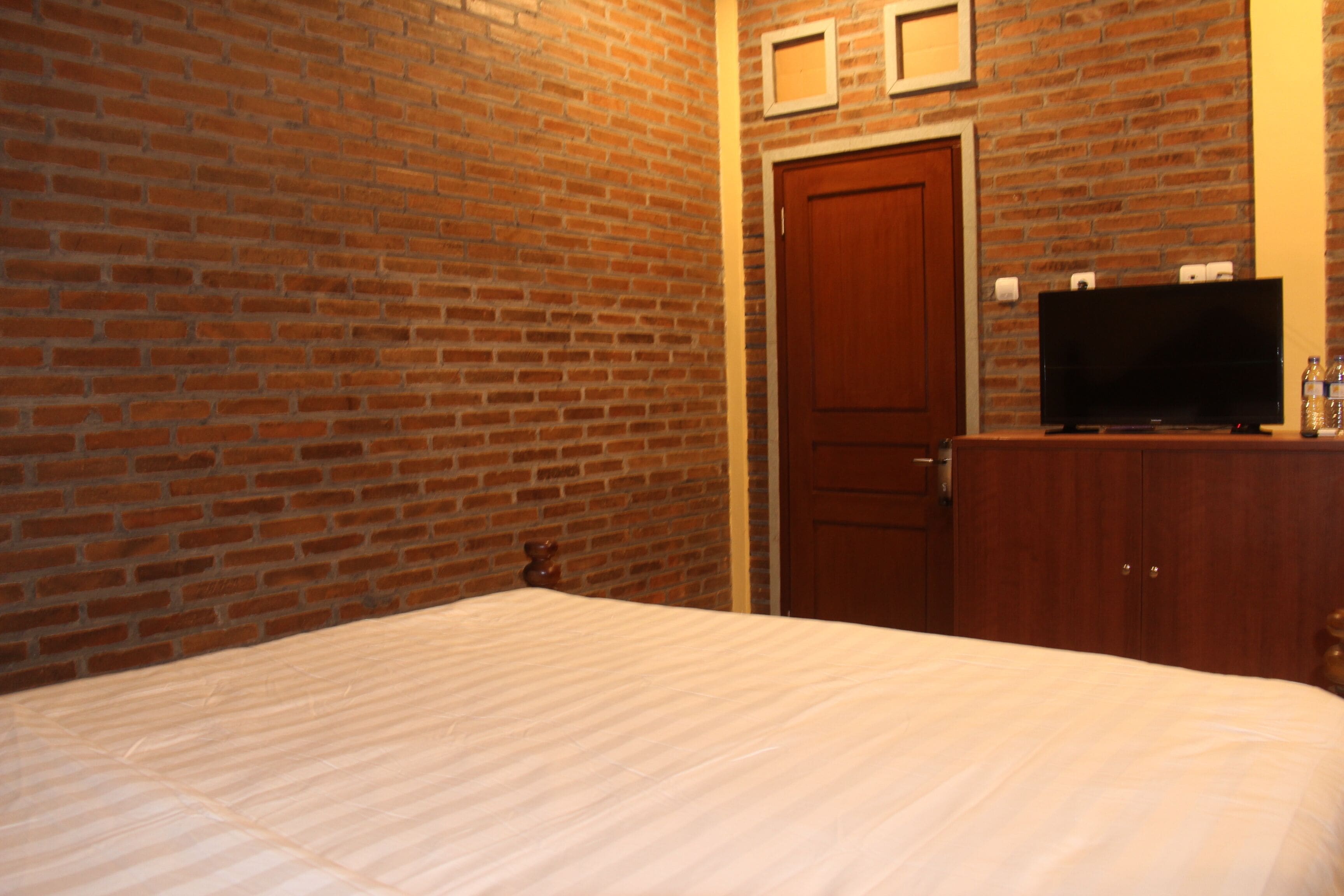 A hotel room with a brick wall, a wooden door, a television on a wooden cabinet, and a bed with a white bedspread.