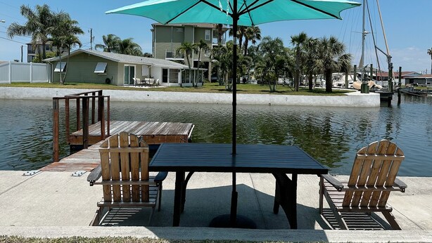 A waterfront patio with a table and chairs, a large umbrella, and a view of the water and buildings in the background.