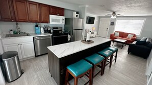 A modern kitchen with a central island, bar stools, a dining area with a sofa, and a living room with a TV.