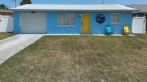 A single-story building with a blue exterior, a yellow door, and a garage. There are two Adirondack chairs outside.