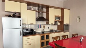 Cozinha compacta com geladeira, fogão, micro-ondas e armários de madeira. Há uma mesa de jantar com cadeiras e piso estampado.