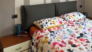 Uma cama com colcha florida colorida e duas almofadas, uma mesa de cabeceira de madeira com uma caneca azul, e uma parede com listras verticais.