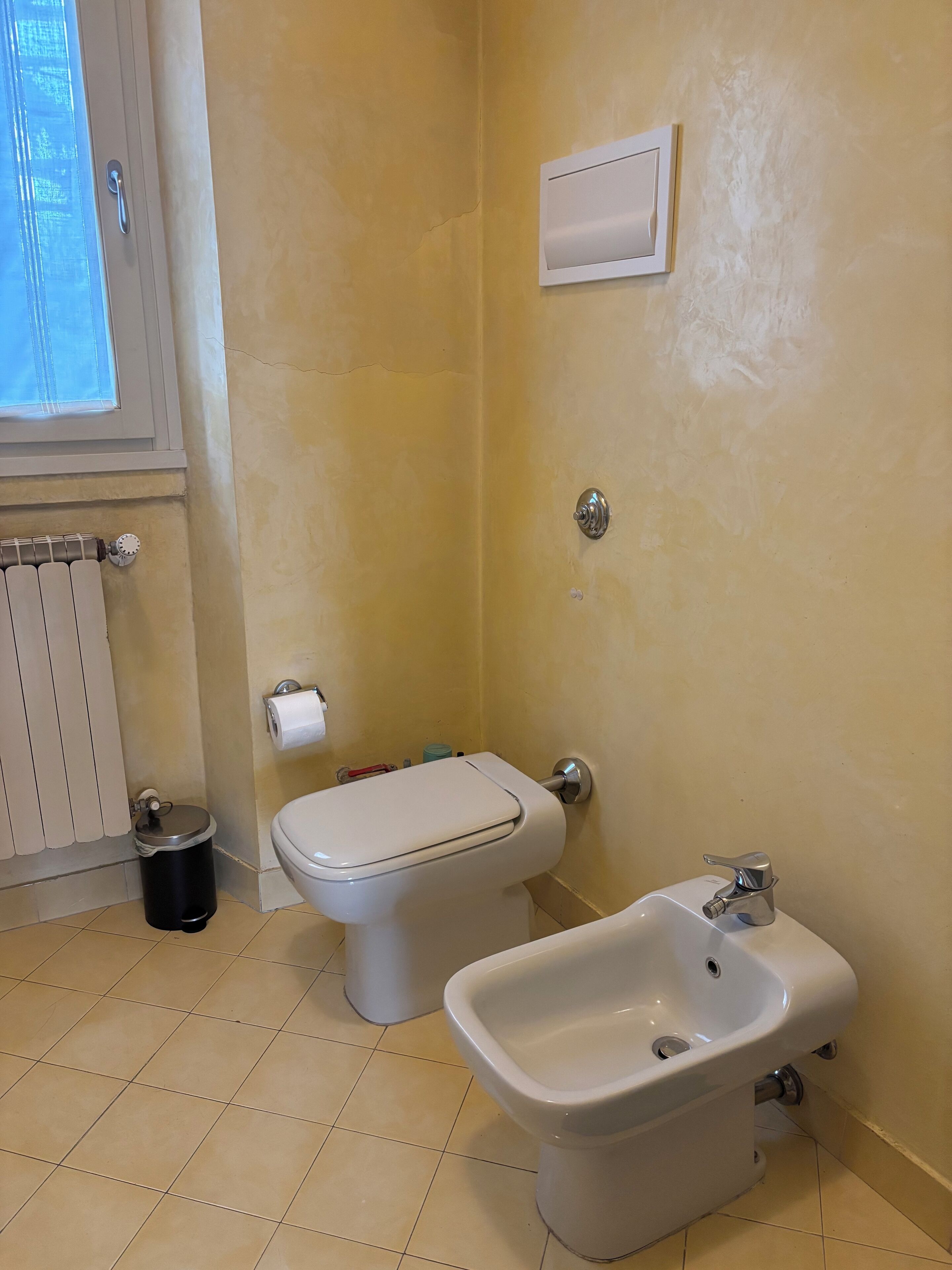 A bathroom with a toilet, bidet, radiator, and a window.