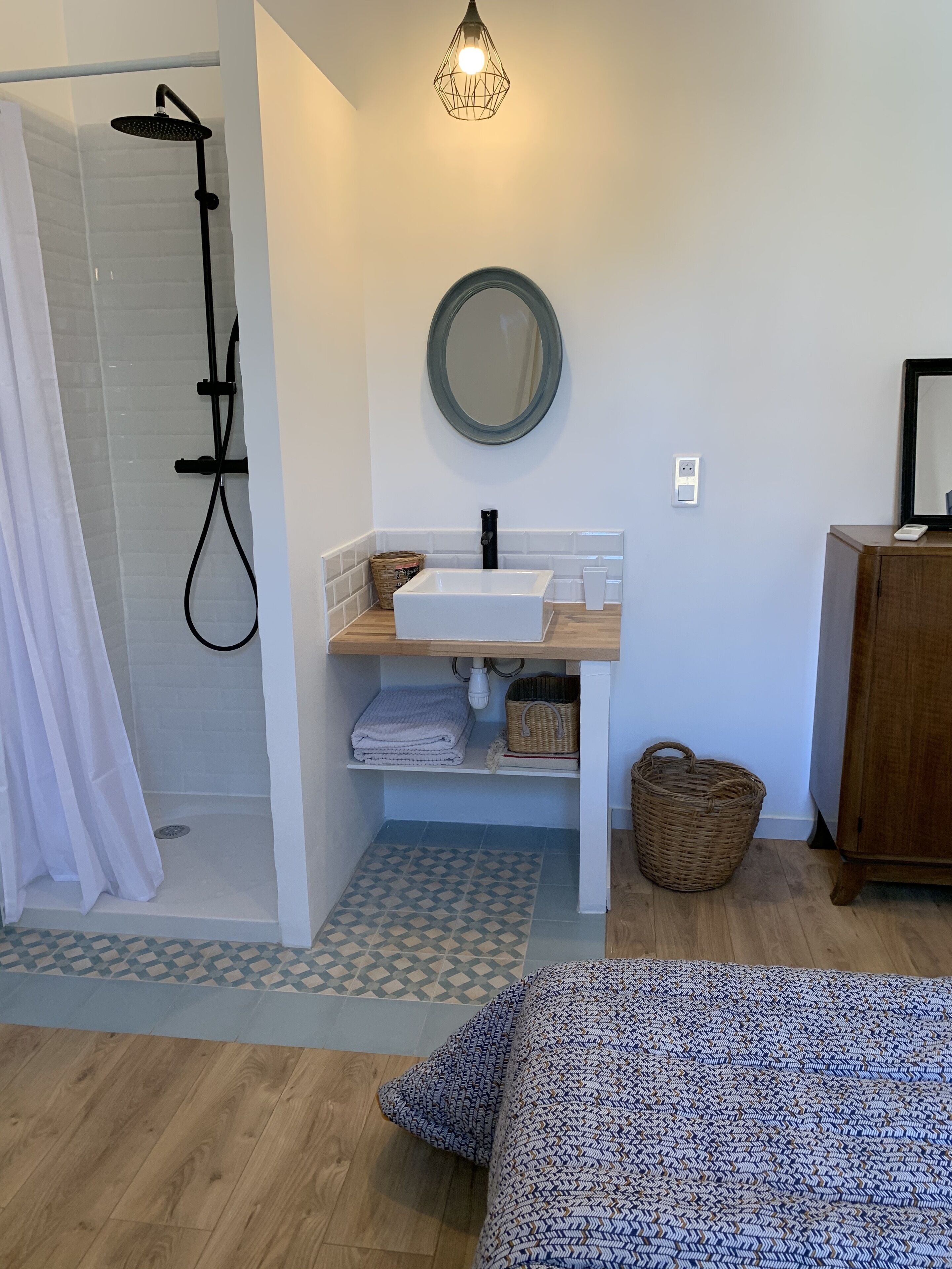 Ein Badezimmer mit Dusche, Waschbecken und Spiegel. Ein Holzschrank und ein Bett mit gemustertem Überwurf sind ebenfalls vorhanden.